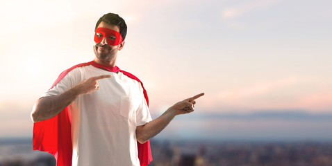 Superhero man with mask and red cape pointing to the lateral on a sunset background © luismolinero