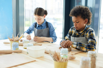 Diligent schoolkids in workwear kneading clay at lesson in workshop