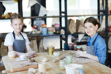 Fototapeta premium Two youthful schoolgirls looking at you while siting by table and kneading clay to make handcraft