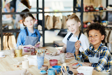 Intercultural schoolkids sitting by big table, looking at camera and painting handmade clay mugs