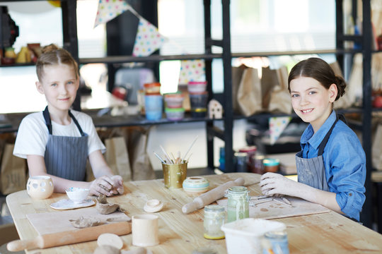 Two youthful schoolgirls looking at you while siting by table and kneading clay to make handcraft