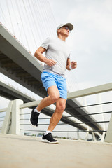Obraz premium Aged man in sportswear running along bridge in urban environment on summer morning