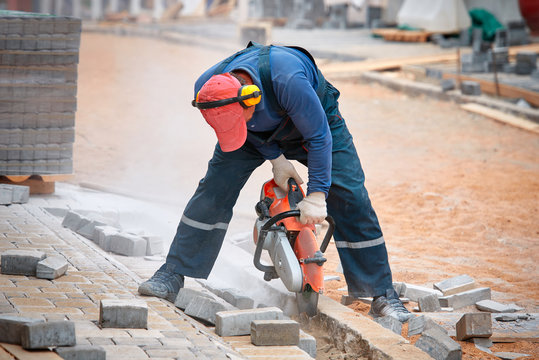 Construction Worker Cuts Walkway Curb With Circular Saw. Man Protect Hearing From Noise Hazards On The Job. Tiles Piled In Pallets On Background. Saw Paving Slub Next To The Worker