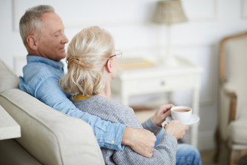 Senior man embracing his wife with cup of tea while both sitting on sofa in living-room