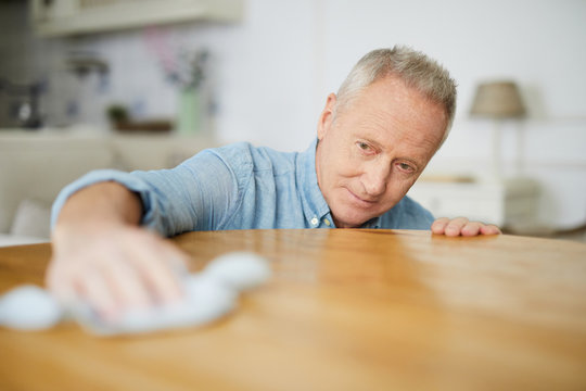 Senior man leaning over table while wiping it with duster during housework