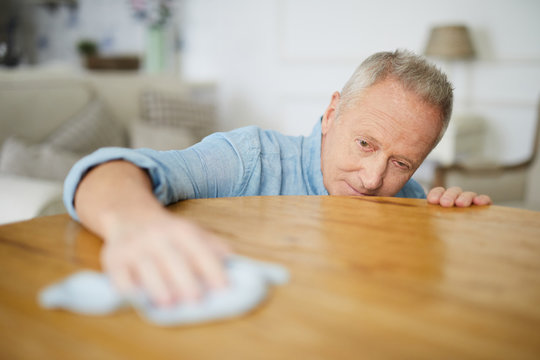 Aged man rubbing wooden table in living room with duster while carrying out domestic chores