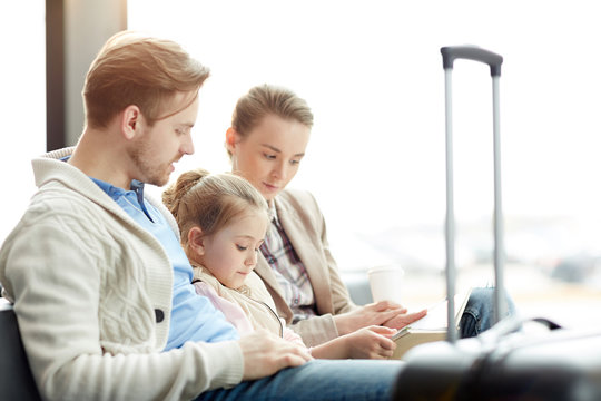 Young Family Of Three Sitting In Airport Lounge Before Airplane Departure And Reading Book