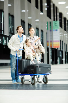 Little girl pointing at timetable with departure data while sitting on baggage cart with her mother and father near by