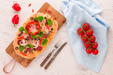 Homemade pizza with ham, pepperoni, mushrooms, capers and olives on a white concrete surface.