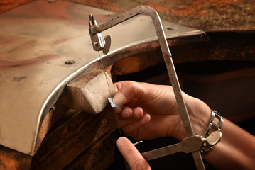 hands of a female goldsmith work on a piece of silver with a metal saw on the workbench, close up