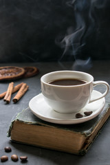 A cup of coffee with steam on an old book, cinnamon sticks and oranges in the background