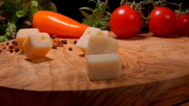 Cubes of Parmesan cheese fall to the wooden board on the background of greenery and vegetables