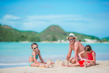 Fototapeta premium Family making sand castle at tropical white beach. Father and two girls playing with sand on tropical beach