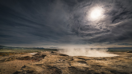 Strokkur - Geysir - island - gleissende Sonne direkt über angestrahltem Geysirloch mit emporsteigendem Dampf und dramatischem Himmel