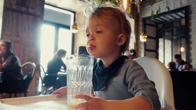 Baby boy drinks orange juice glass sitting at table in cafe