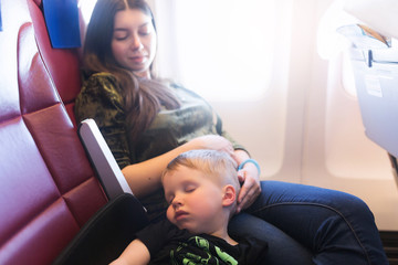 Young mother with sleeping 3 years old baby sitting together in airplane cabin near window. Child sleeps during the flight by plane