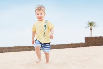 Little happy child boy with blue eyes and joyful smile runs on tropical beach. Three years old kid...