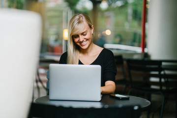Portrait of a young and attractive blonde girl working on her notebook laptop inside a cafe. She is sitting and smiling as she focuses on finishing her work.