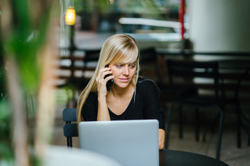 A good-looking blonde woman is talking on her smartphone as she takes a break from working on her laptop in a cafe.