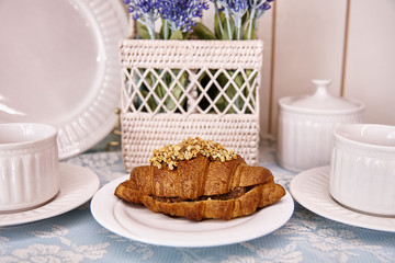 Croissant with a filling on a white plate next to white tableware and a basket of flowers.