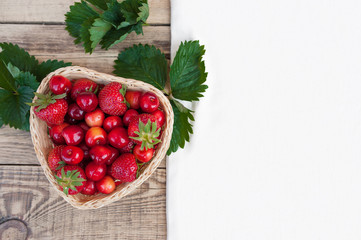 Yellow-orange fresh cherries and red ripe strawberries on a white plate, on a wooden light table with a texture, with green leaves and a white tablecloth and napkin.