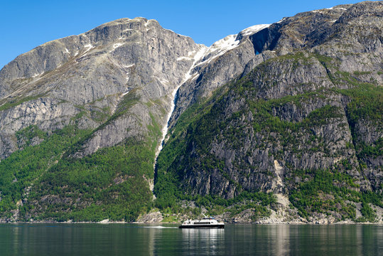 Small tour boat or ferry in a fjord on a sunny day. Mountain peaks with snow and ice in the background. Location Eidfjord in Hordaland, Norway.