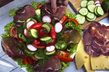 A hand of a cook making salad of fresh vegetables with bacon; set of delicious healthy food products: sliced cheese and meat, cucumbers, radish, sweet pepper, tomatoes and lettuce.