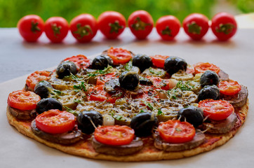 Veggie pizza with mushrooms, black olives and herbs on the gray kitchen table decorated with fresh cherry tomatoes. Tasty pizza on the green blurred background. Side view