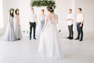 Beautiful bride in a white dress holds a wedding bouquet and is going to her groom with bridesmaids and groommen for wedding ceremony. White room is decorated with pine, flowers and blue candles.