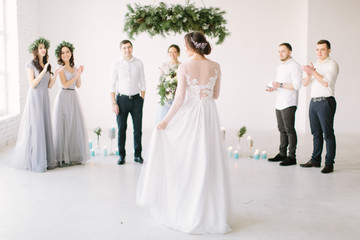 Beautiful bride in a white dress holds a wedding bouquet and is going to her groom with bridesmaids and groommen for wedding ceremony. White room is decorated with pine, flowers and blue candles.