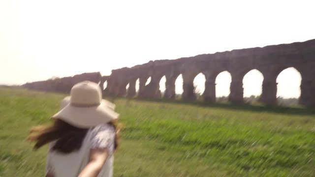 Come with me. Young beautiful girl with hat leading man boyfriend holding hands toward roman aqueduct arches in parco degli acquedotti park ruins in rome romantic lovely happy couple slow motion
