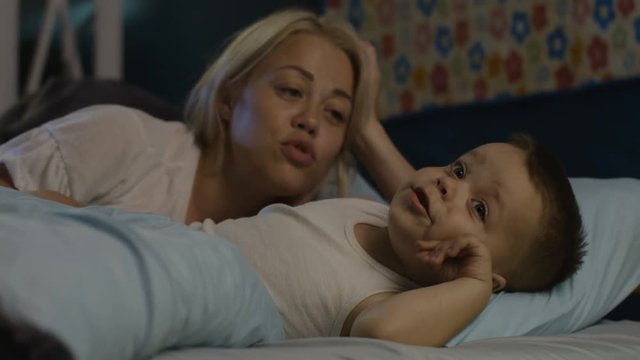 Beautiful woman lying with little boy in bed and telling interesting story before going to sleep at night