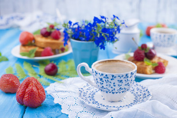 Small cupcakes with berries, strawberries and cherries. Delicious sweet breakfast and fragrant coffee, on a blue wooden background and flowers. Copy space