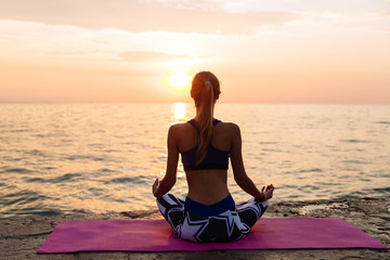 Healthy concept. View from back of sportive woman meditating in lotus pose, sitting on the pier, looking at sea sunset, enjoying the yoga.