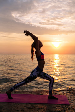 Yoga at sunrise. Prifile of attractive woman doing exercises for body, practicing yoga, standing against the sunrise sea background. On pier. Dressed in leggings and singlet.