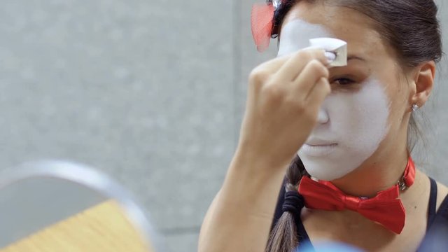 Pretty young mime applying white paint on her face in front of small mirror