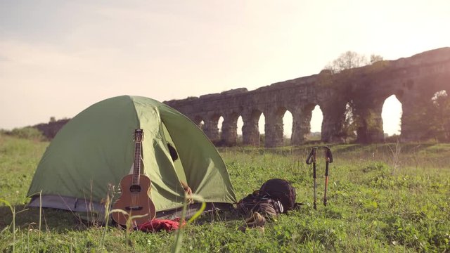 Young man backpacker wakes up exits camping tent in front of roman aqueduct arches in parco degli acquedotti park ruins in rome at sunset sticks in ground slow motion