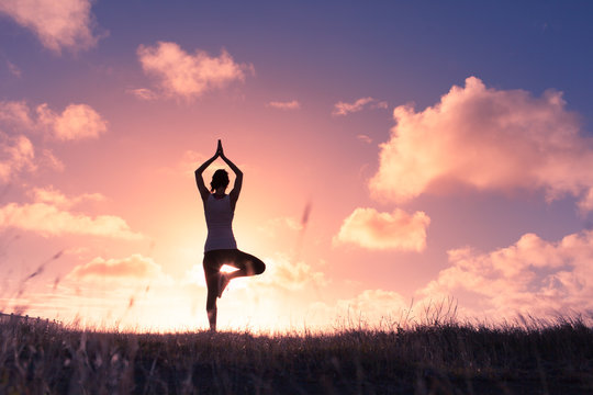 Young female doing balancing yoga pose against colorful sunset. 
