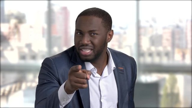 Black businessman humiliating someone. Annoyed displeased dark-skinned man in business suit accusing someone and pointing with finger on blurred background. Human negative expressions.