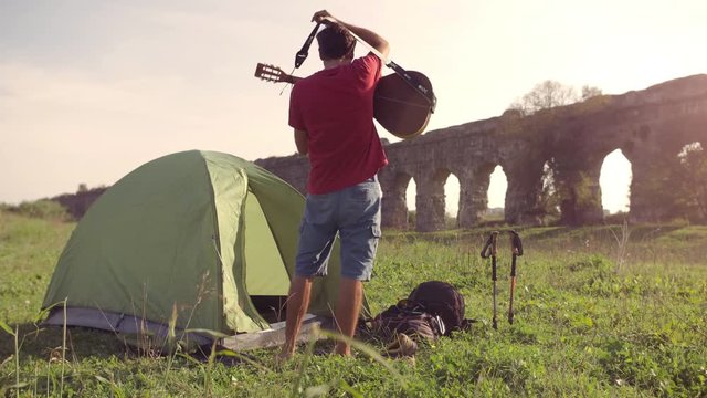 Young man backpacker outside camping tent picks up guitar and walks on grass in front of roman aqueduct arches in parco degli acquedotti park ruins in rome at sunset sticks in ground slow motion
