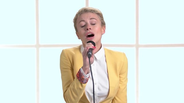 Young pretty blonde singing with microphone. Charming girl is singing on abstract window background. Beautiful caucasian performer.