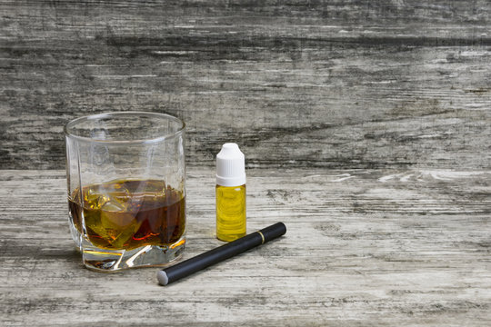Whiskey in a glass with an e-cigarette and liquid for it on a gray stone background 
 