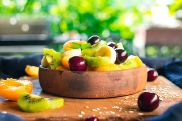 Fruit salad in bowl on wooden board. Healthy sweet dessert for breakfast with rip banana, apricot slices, fresh kiwi, cherry berries, mint leaves, cream. Raw vegan vegetarian healthy food