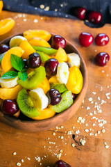 Fruit salad in bowl on wooden board. Healthy sweet dessert for breakfast with rip banana, apricot slices, fresh kiwi, cherry berries, mint leaves, cream. Raw vegan vegetarian healthy food