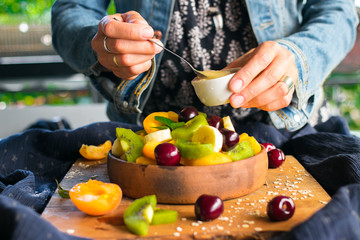 Woman hands adds sweet banana smooth cream with spoon to fruit salad in bowl on wooden board. Healthy sweet dessert for breakfast. Raw vegan vegetarian healthy food