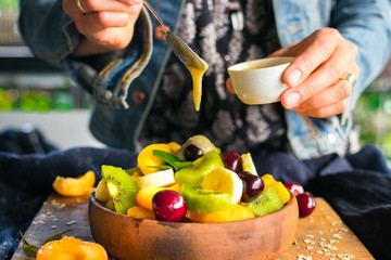 Woman hands adds sweet banana smooth cream with spoon to fruit salad in bowl on wooden board....