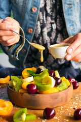 Woman hands adds sweet banana smooth cream with spoon to fruit salad in bowl on wooden board. Healthy sweet dessert for breakfast. Raw vegan vegetarian healthy food