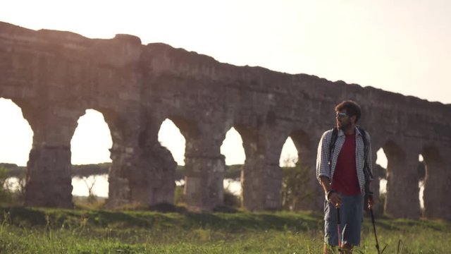 Young man backpacker walks barefeet with sticks on grass in front of roman aqueduct arches in parco degli acquedotti park ruins in rome at sunset slow motion