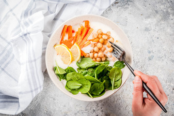 Modern vegan food, person eat  savory yogurt bowl with beans, chickpeas, spinach, spicy carrots, lemon, Gray stone background, copy space top view, hands in picture