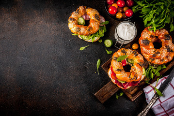 Variety of homemade bagels sandwiches with sesame and poppy seeds, cream cheese,  ham, radish, arugula, cherry tomatoes, cucumbers, with ingredients on dark concrete background copy space top view
